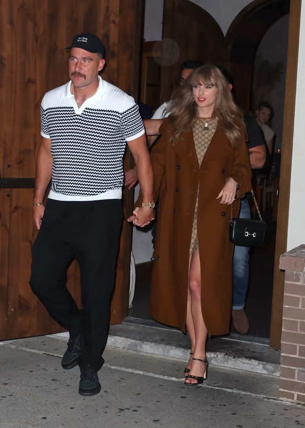 Homem com boné preto, camisa polo listrada em preto e branco e calça preta. Mulher com longo casaco marrom, vestido bege com estampa, sandálias de salto alto e bolsa preta clássica. Estilo casual sofisticado e tendências de moda outono/inverno.