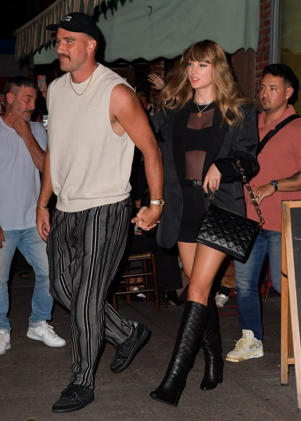 Casal trendy em evento noturno: homem usa colete branco sem mangas, correntinha dourada, calça listrada e tênis preto. Mulher com blazer oversized preto, blusa transparente, mini saia preta, botas de cano alto e bolsa de couro preta.