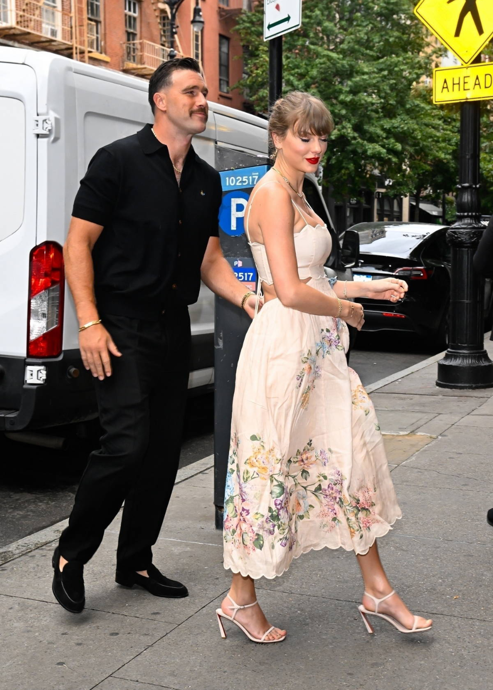 Mulher vestindo um vestido midi de alcinha, floral, creme e sandálias de salto fino bege. Homem com camisa preta, calça preta e sapatos pretos. Estilo casual e elegante, combinando tendências de moda urbana e romântica.