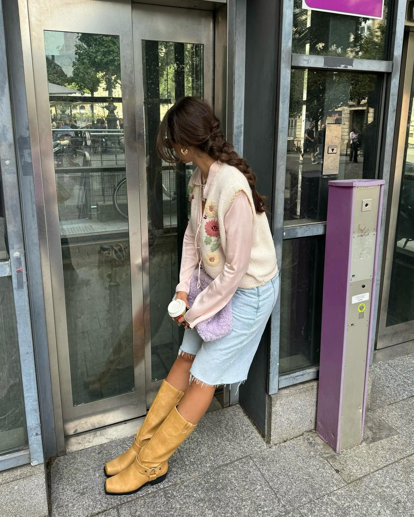 Mulher com visual casual e feminino. Usa colete com bordado floral, camiseta de manga longa, saia jeans desfiada e botas de cano alto bege. Segura um copo de café e uma bolsa de pelúcia lilás. Estilo retrô com toque moderno.