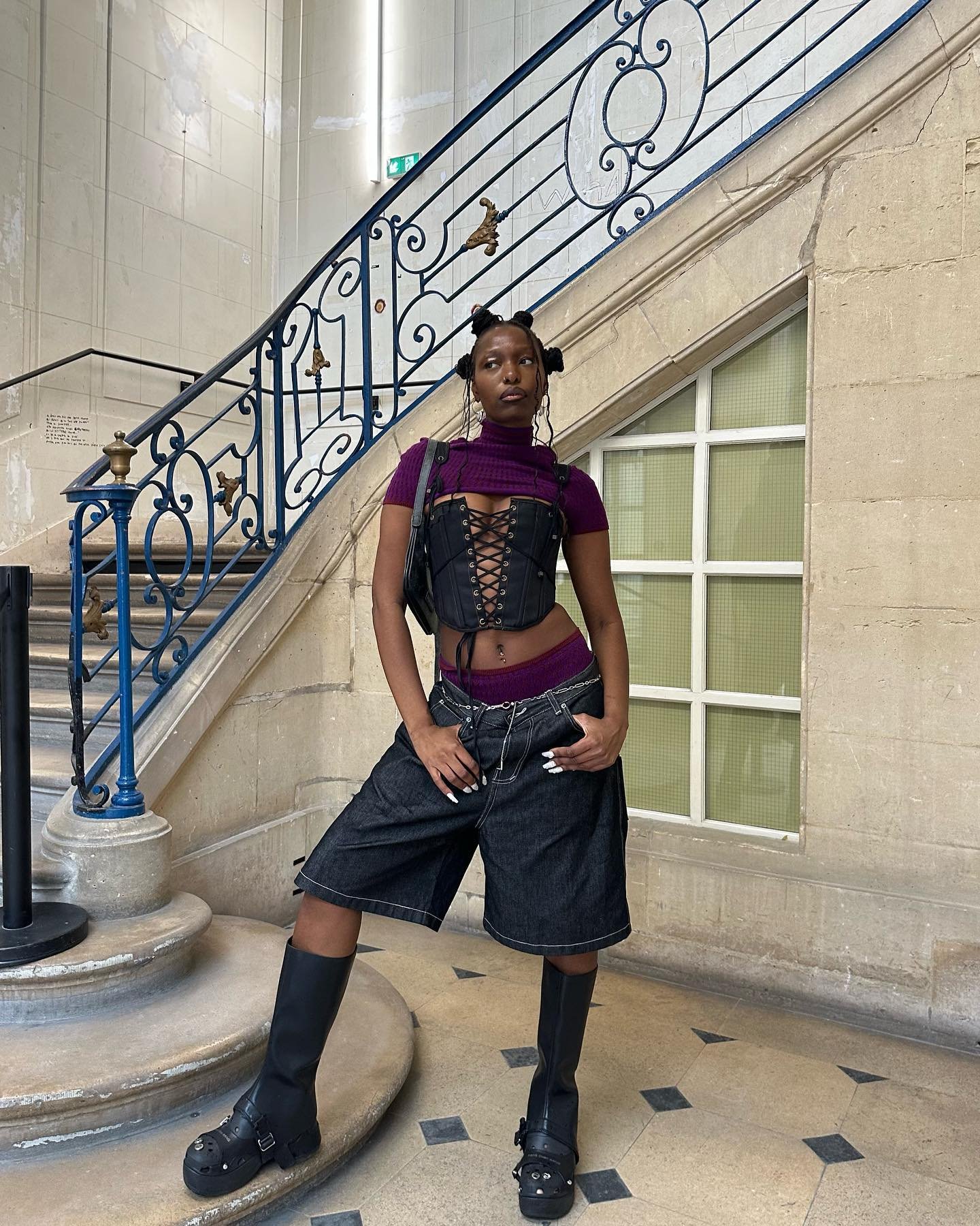 Jovem vestindo top cropped roxo, espartilho preto, bermuda jeans larga, meias de cintura alta e botas pretas robustas. O estilo mistura elementos góticos e streetwear, destacando tendências contemporâneas e ousadas na moda urbana.