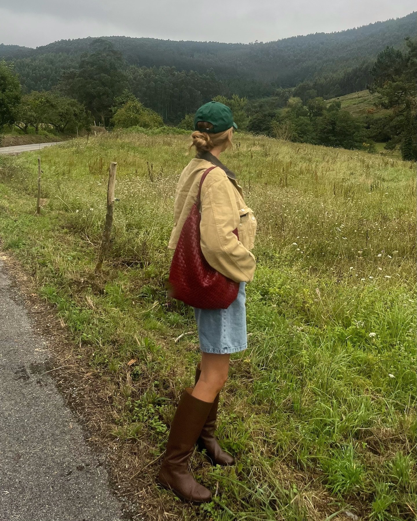 Pessoa vestindo jaqueta bege, boné verde, short jeans, botas marrons de cano alto e bolsa grande vermelha, em um ambiente rural. O look mistura casual com elementos rústicos e confortáveis, ideal para passeios ao ar livre em dias frios.