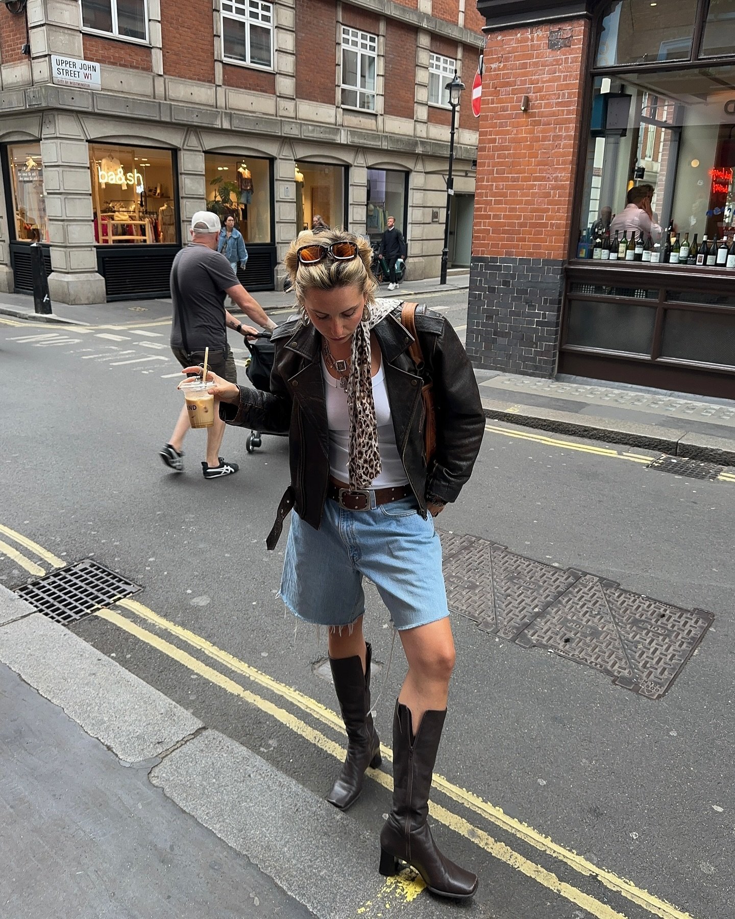 Mulher com look casual estiloso: jaqueta de couro preta, blusa branca, shorts jeans desfiados, cintos marrom, lenço de animal print e botas pretas de cano alto. Ela segura um café gelado e usa óculos de sol no cabelo, criando um visual urbano e moderno.