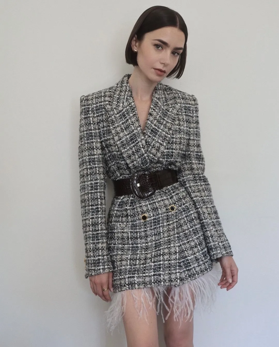 Mulher usando blazer de tweed xadrez cinza, estilo oversized, com cinto preto largo marcando a cintura. Detalhes com penas brancas na barra da peça. Visual sofisticado e moderno, combinando elementos clássicos com tendências contemporâneas.