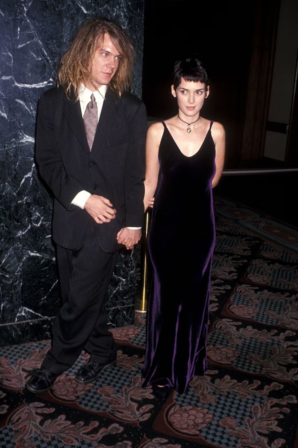 Casal posando: Ele veste terno preto com gravata estampada e camisa branca, com cabelo bagunçado; ela usa vestido longo de veludo preto com alças finas e colar choker, cabelo curto e penteado. Looks da Winona Ryder.