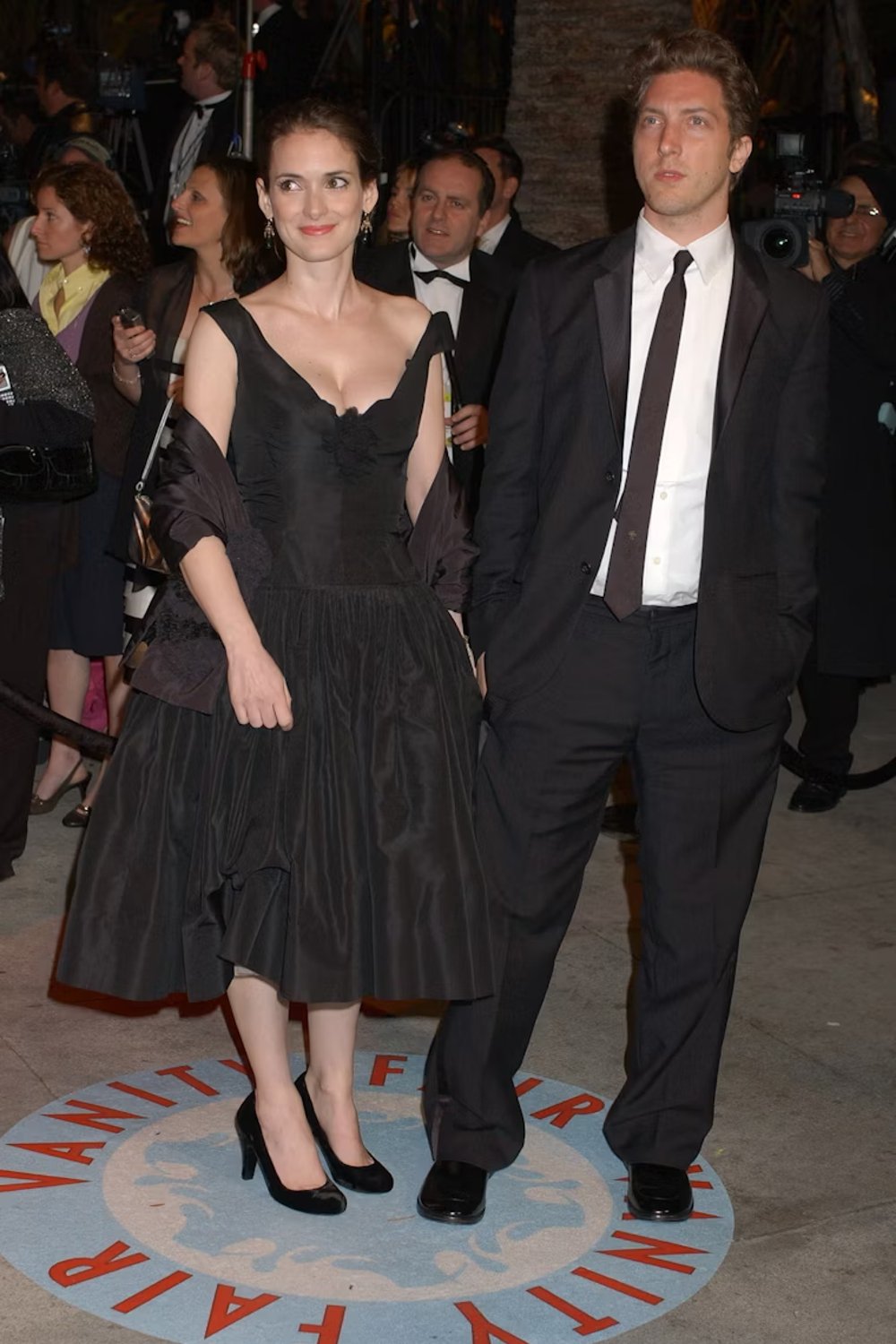 Casal elegante em um evento de gala. Ela veste um vestido preto clássico com decote, sapatos de salto alto e envolve os ombros com um xale preto. Ele usa um terno preto com gravata e camisa branca. Looks da Winona Ryder.