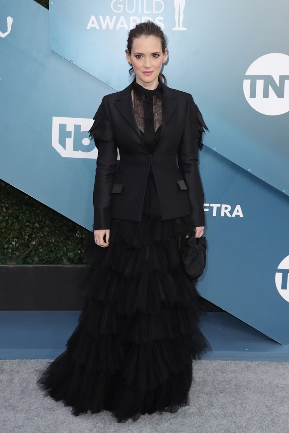 Celebridade em evento de premiação usando vestido preto longo com múltiplas camadas de tule, blazer preto ajustado por cima e camisa transparente. Looks da Winona Ryder.