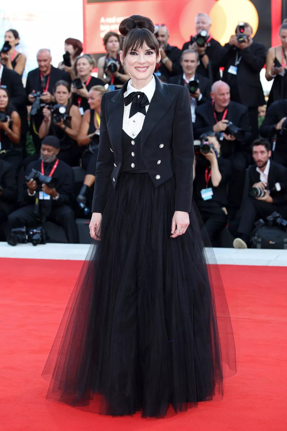 Pessoa em evento com traje estilo smoking feminino, composto por blazer preto com botões metálicos, camisa branca, gravata borboleta preta e saia de tule longa preta. Looks da Winona Ryder.