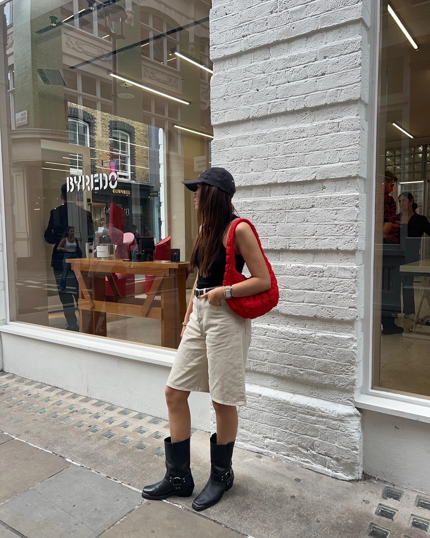 Mulher de cabelo solto, usando shorts bege, top preto, botas pretas e bolsa vermelha acolchoada. Ela também usa boné preto, criando um look casual e urbano. Estilo destaca tendências de mix de acessórios coloridos com peças básicas e confortáveis.