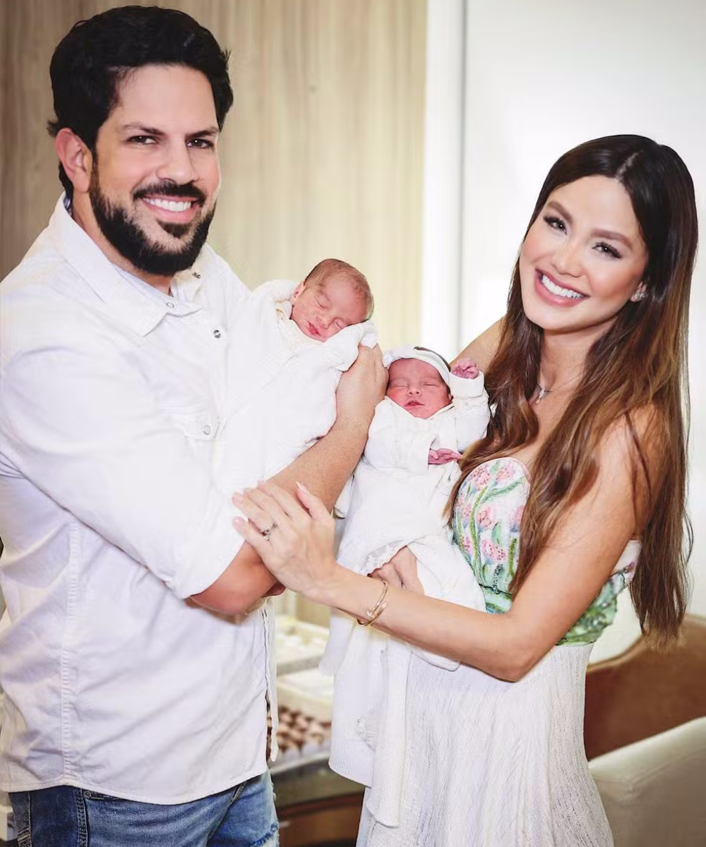 Casal com gêmeos recém-nascidos. Ele veste camisa social branca e jeans, enquanto ela usa vestido longo com corset floral e saia branca fluida. O look dela combina delicadeza e elegância, enquanto o dele é casual e moderno. Os bebês estão envoltos em