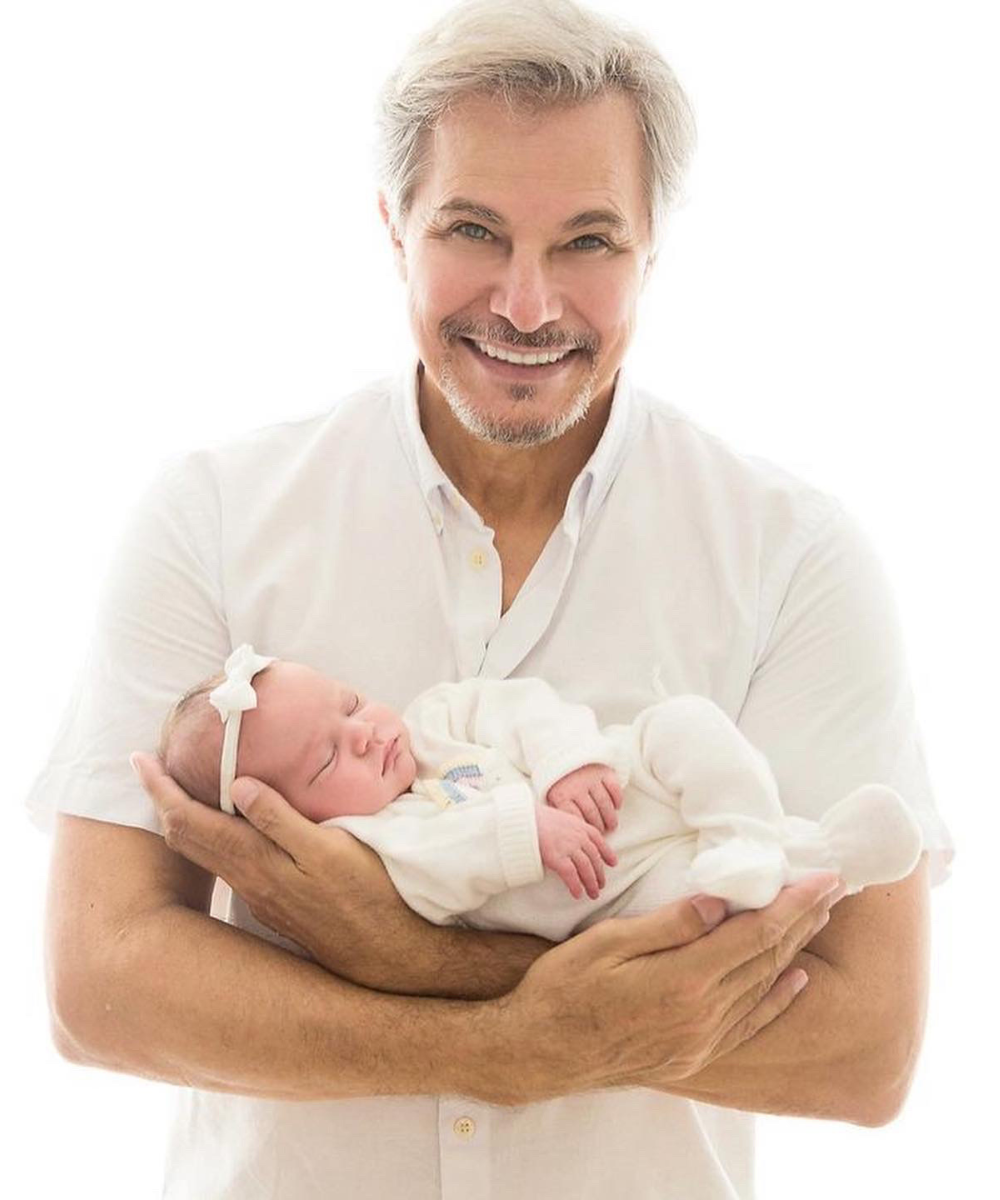 Homem de cabelos grisalhos, vestindo camisa social branca, segurando um bebê recém-nascido, também vestido de branco com tiara de laço. O look casual do homem, com barba bem aparada, e o traje delicado do bebê refletem tendências de moda clássica e clean.