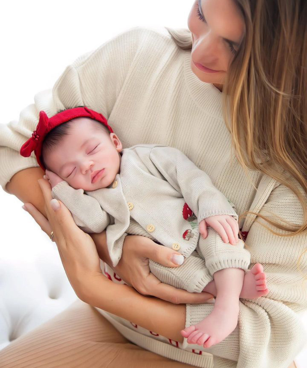 Bebê vestindo conjunto de tricô bege com detalhes florais, combinando com faixa vermelha na cabeça; mulher usando suéter de tricô bege, criando um visual aconchegante e harmonioso. Tendências de moda infantil e roupas confortáveis para mãe e bebê.