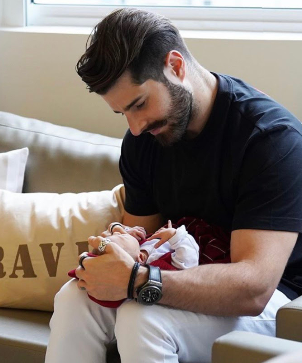 Homem com cabelo estilizado e barba bem cuidada veste camiseta preta e calças claras. Acessórios incluem relógio de pulso e anéis. Ele segura delicadamente um bebê, sugerindo um toque de estilo casual e moderno, combinando conforto e tendência.