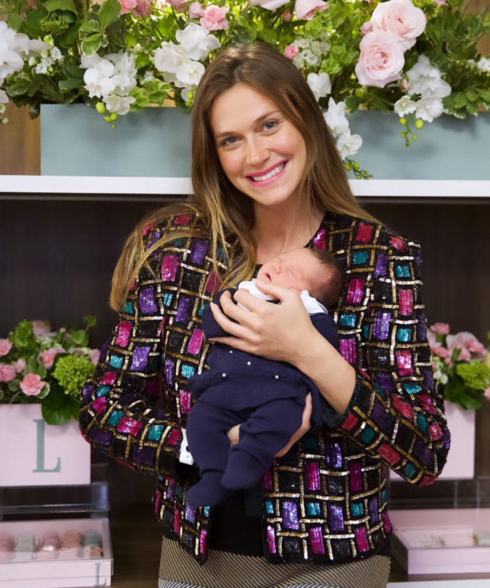 Mulher sorridente segura um bebê descansando em seus braços. Ela veste uma jaqueta colorida e brilhante com padrões geométricos em roxo, rosa, azul e dourado. Ao fundo, há arranjos florais delicados e caixas decorativas.