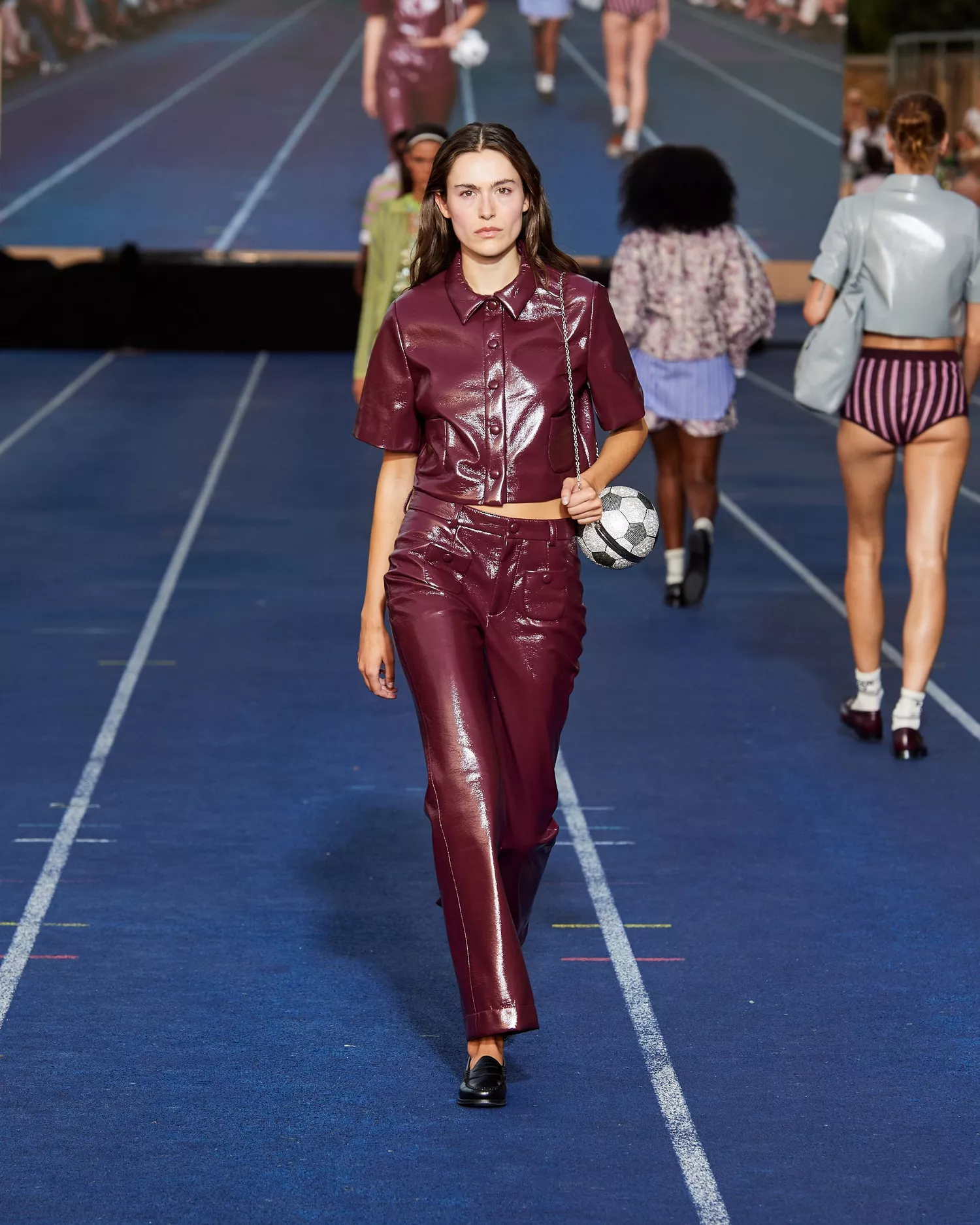 Modelo desfilando com conjunto de couro vinílico na cor vinho, composto por camisa de manga curta e calça. O visual é complementado por bolsa em formato de bola de futebol e sapatos pretos. A peça destaca tendências futuristas e ousadas na moda.