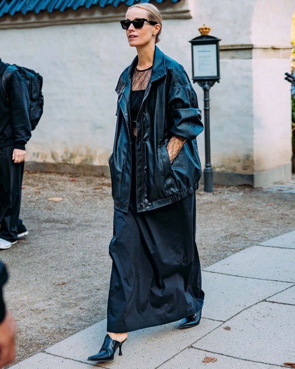 Pessoa usando um vestido preto longo e uma jaqueta de couro oversized preta, combinados com sapatos de salto. O look é completado com óculos de sol estilo retrô.