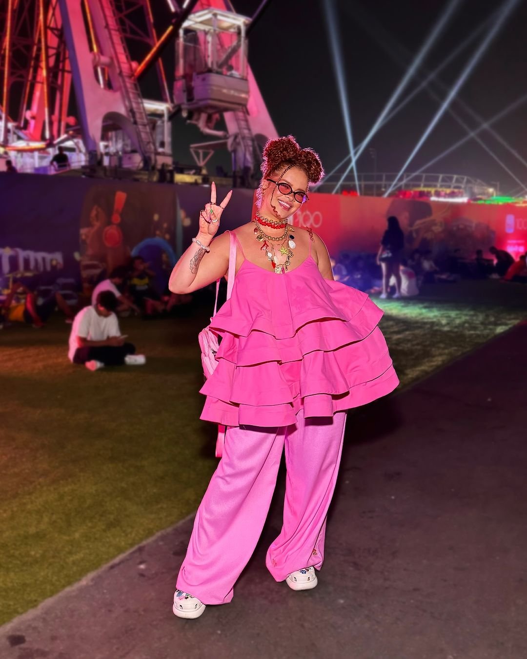 Pessoa em festival noturno usando look monocromático rosa vibrante, composto por blusa com babados, calça larga e tênis. Acessórios incluem colar colorido e óculos de aro grande, refletindo estilo ousado e alegre. Looks Rock in Rio 2024.