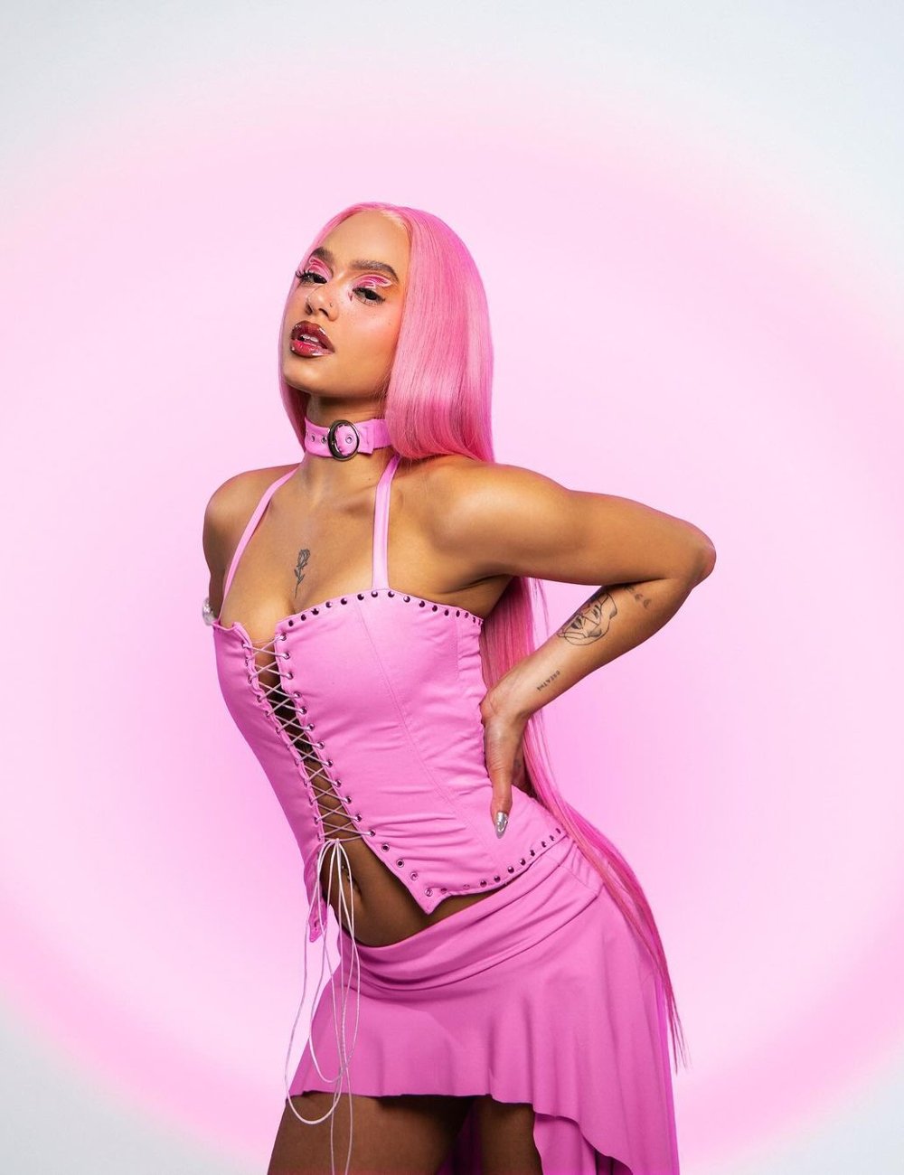 Imagem de uma mulher com cabelo rosa longo, vestindo um corset rosa com amarração frontal e saia assimétrica combinando. Completa o look com maquiagem artística e gargantilha rosa. Looks Rock in Rio 2024.