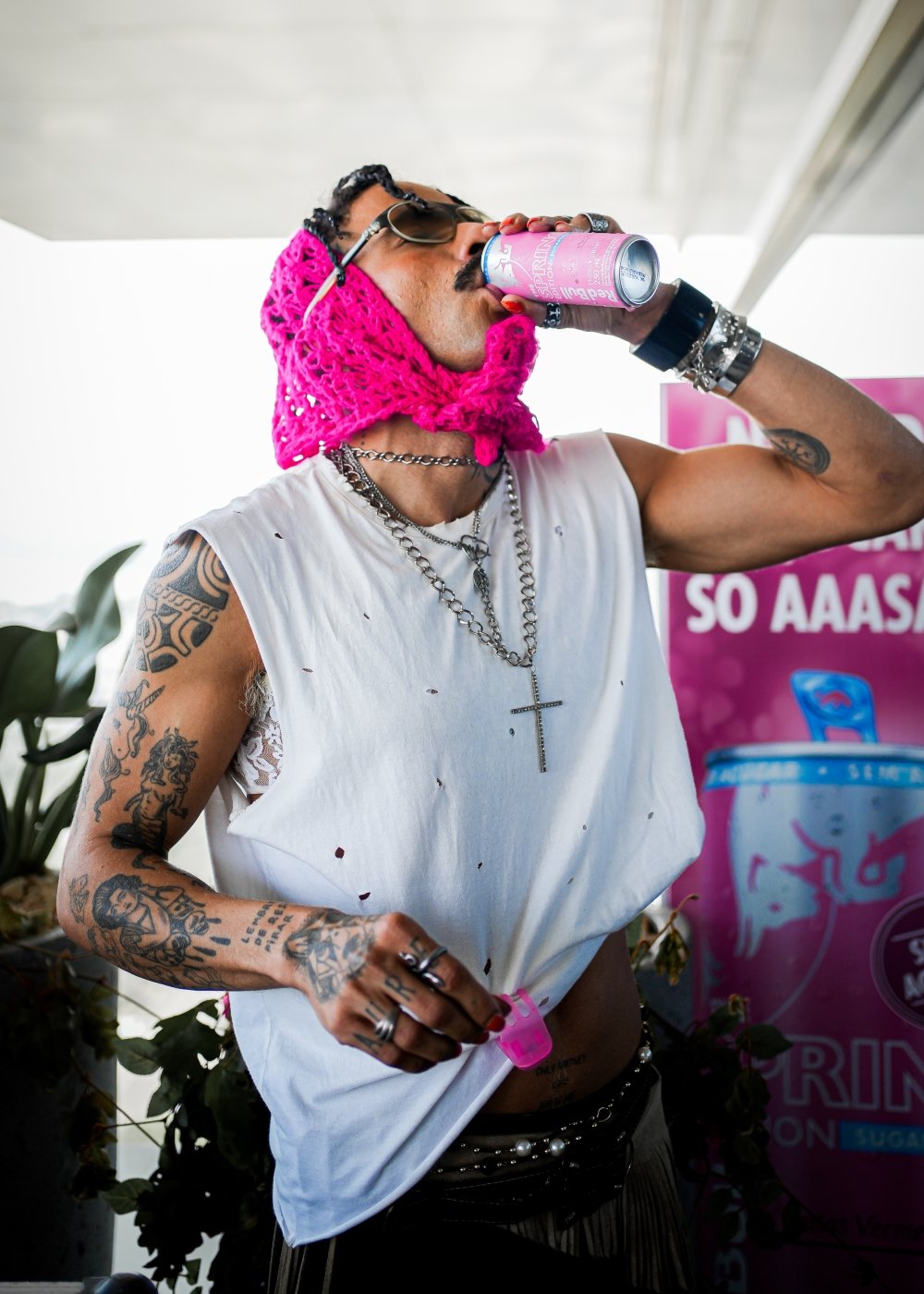 Homem usando toca de crochê rosa, camiseta branca rasgada, colares de corrente, pulseiras prateadas e anéis. Visual com influências punk e alternativas, destacando tatuagens visíveis nos braços. Looks Rock in Rio 2024.