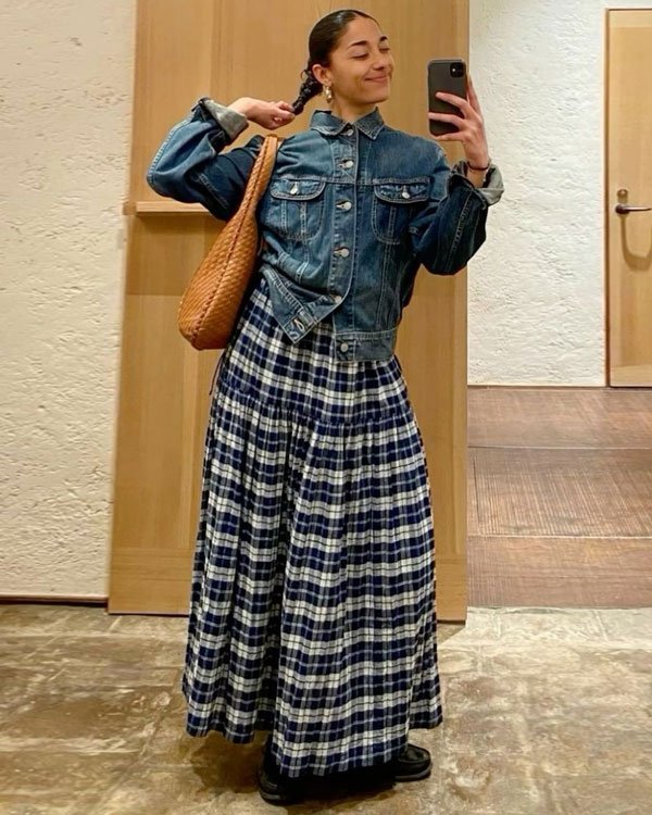 Mulher vestindo jaqueta jeans, saia longa xadrez em tons de azul e branco, e calça botinas pretas. Bolsa grande de palha marrom no ombro. Look casual e confortável, combinando tendências vintage e retrô com um toque moderno.