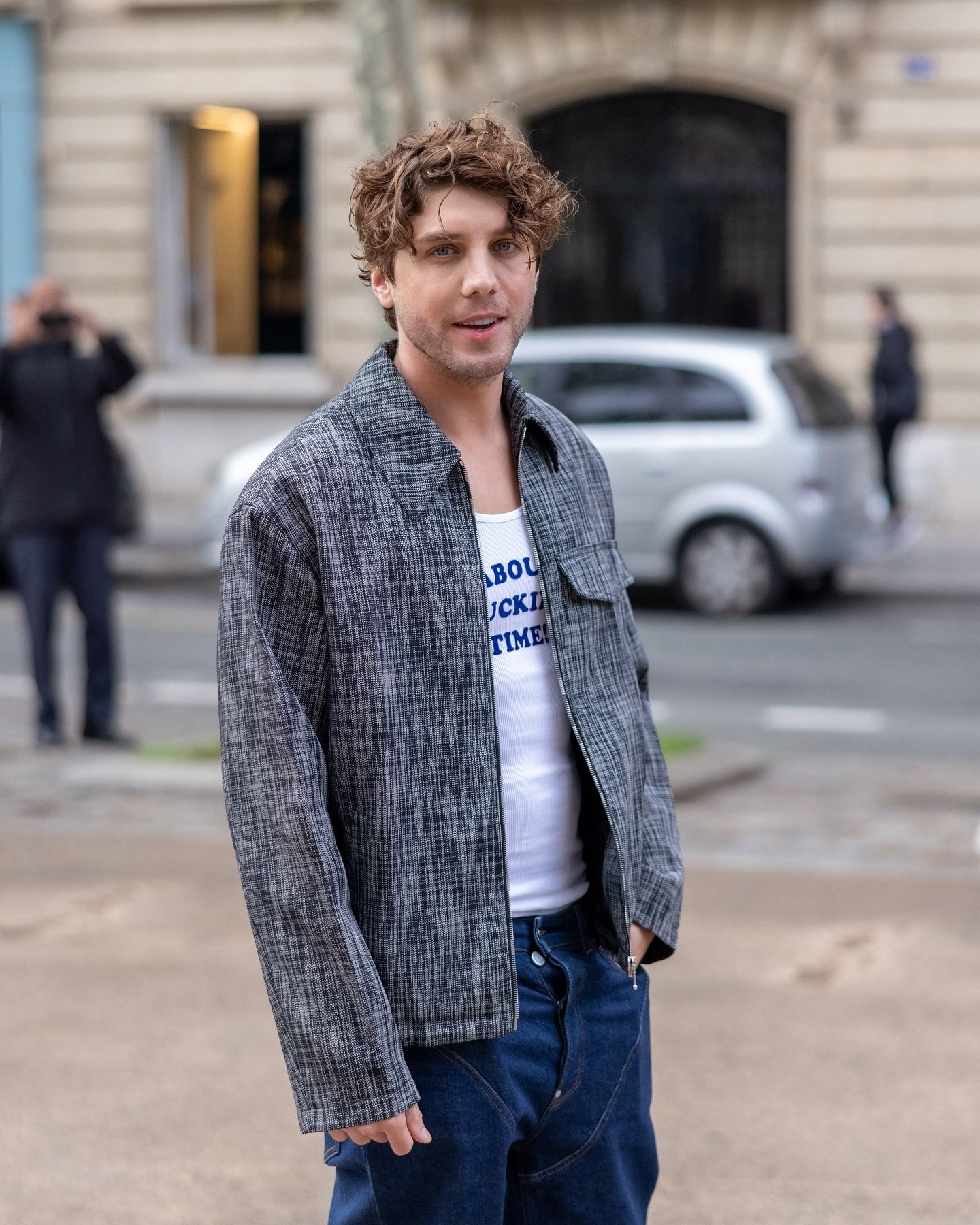 Homem com cabelo ondulado e despojado, usando jaqueta xadrez clássica sobre camiseta branca com slogan azul, calça jeans folgada. Look casual misturando tendências modernas e retrô, ideal para ocasiões informais e estilos urbanos.