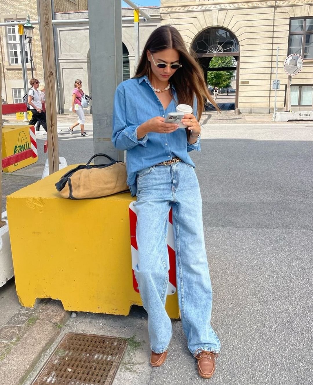 Mulher em look casual urbano, com camisa jeans oversized e calça jeans de corte largo, combinados com sapatos marrons e óculos de sol estilo retrô. Acessórios incluem colar de pérolas e bolsa casual.