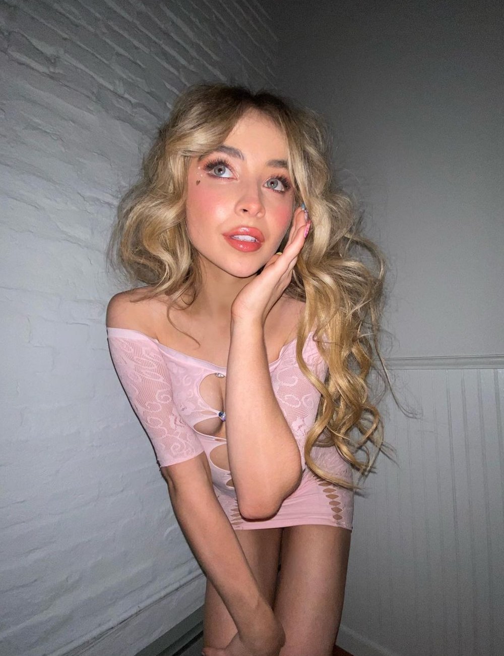 Jovem mulher loira vestindo um vestido curto rosa de ombro caído com detalhes de recortes e amarrações na frente. O visual é complementado por uma maquiagem suave com toque de iluminador e batom nude. Maquiagem da Sabrina Carpenter.