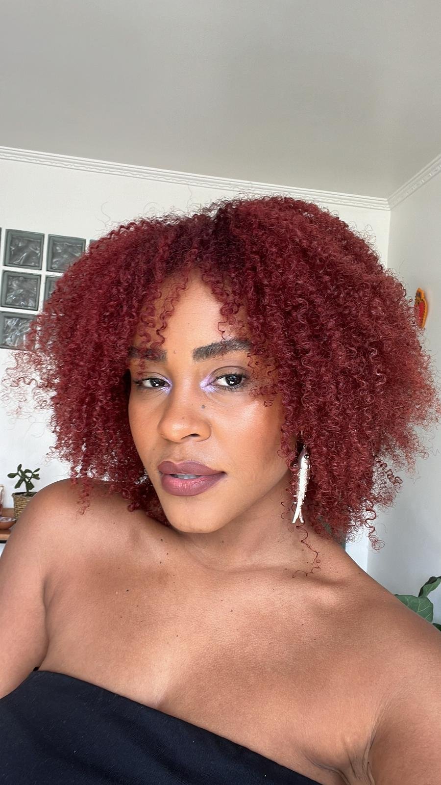 Mulher com cabelo cacheado tingido de vermelho, maquiagem leve com sombra lilás, batom nude e brinco de pendente branco. Está usando um top sem alças preto. O look destaca-se pela combinação de simplicidade e ousadia nas cores e acessórios.