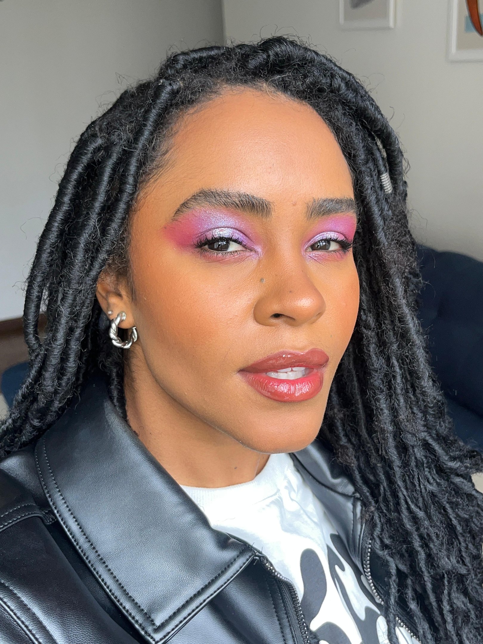 Pessoa com cabelo em estilo dreadlocks, maquiagem vibrante com sombra roxa e blush rosa. Usa jaqueta de couro preta e camiseta branca, tendências comuns na moda urbana e casual. Spets destacam-se com brincos de aro prateados.