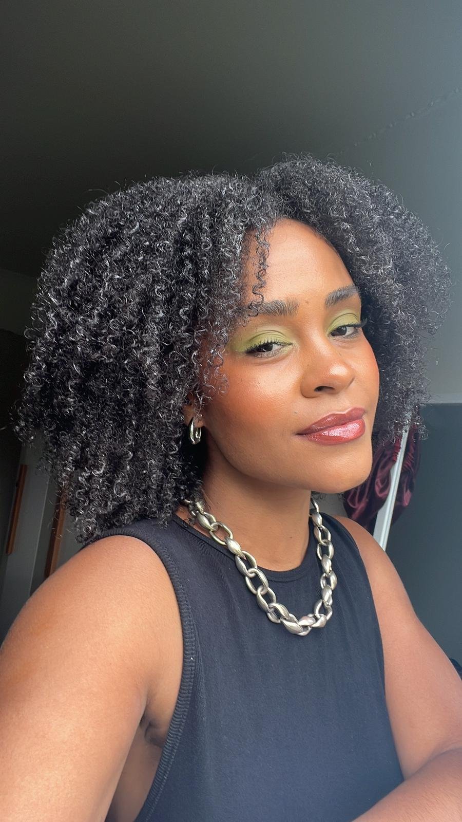 Mulher usando top preto sem mangas e corrente de elos grossos prateada. Maquiagem com sombra verde e batom nude, destacando visual natural e sofisticado. Cabelo cacheado volumoso, tendência em moda afro e empoderamento.