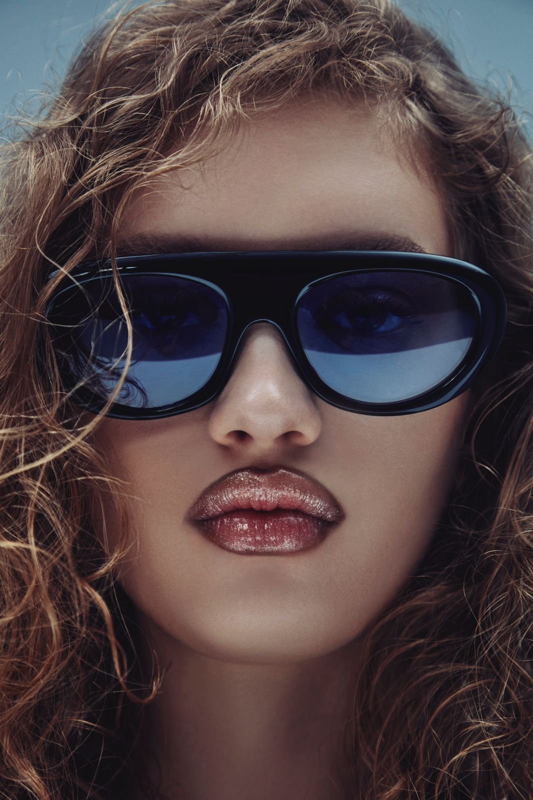 Close-up de modelo com cabelos ondulados, usando óculos de sol oversized de armação preta e lentes escuras. Lágrima acentuada com brilho labial metálico. Estética contemporânea com toque glamouroso. marca de óculos