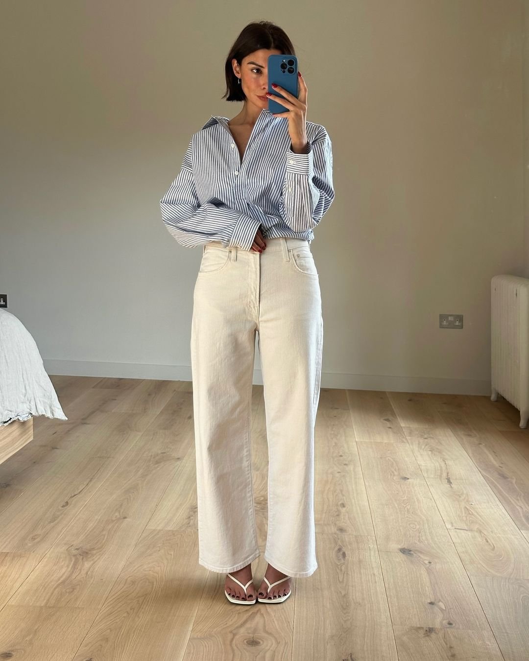 Look casual e minimalista com camisa listrada azul oversized, calça branca de corte reto e sandálias brancas com tiras finas. Tendência de moda com foco no conforto e elegância simples.