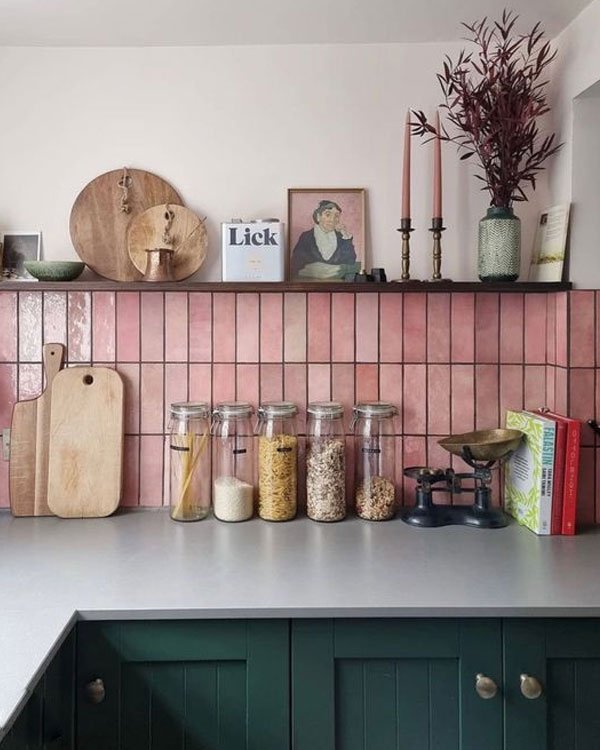 Cozinha moderna e acolhedora com azulejos rosa, armários verdes e decoração minimalista. Prateleira com utensílios e mantimentos em potes de vidro, retratando uma tendência de organização e estilo rústico-chic.