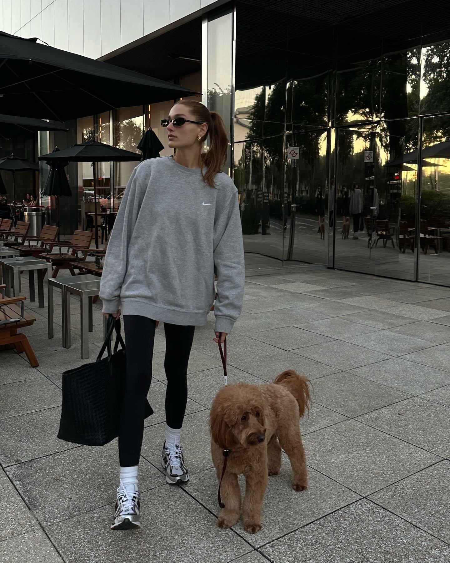 Pessoa usando um moletom cinza, legging preta e tênis esportivo. Acessórios incluem óculos de sol escuros, brincos pequenos e bolsa preta. Look casual, confortável e moderno, ideal para um passeio com cachorro em ambiente urbano.