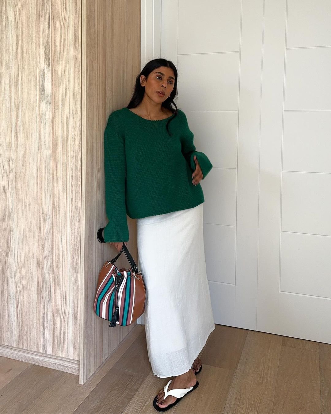 Mulher usando suéter verde oversized e saia branca longa. Completa o look com sandálias brancas e bolsa de ombro listrada, estilo casual e confortável. Elementos-chave: suéter oversized, saia longa, sandálias comfort, bolsa listrada.