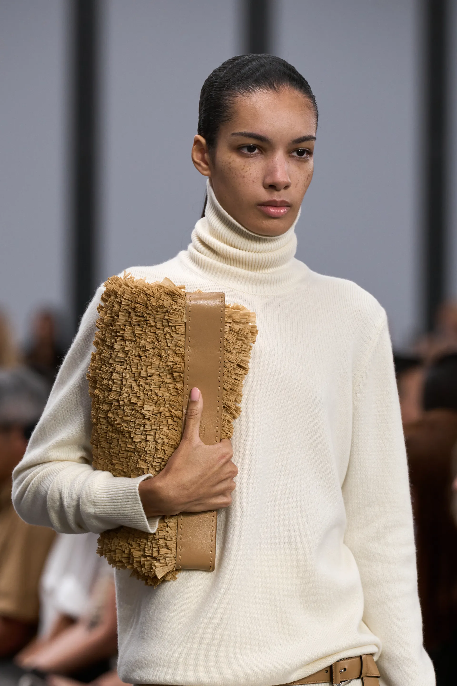 Modelo usando suéter off-white de gola alta em look minimalista, segurando clutch texturizada bege. Tendência outono/inverno 2023, combina tons neutros e peças básicas sofisticadas. NYFW 2025.