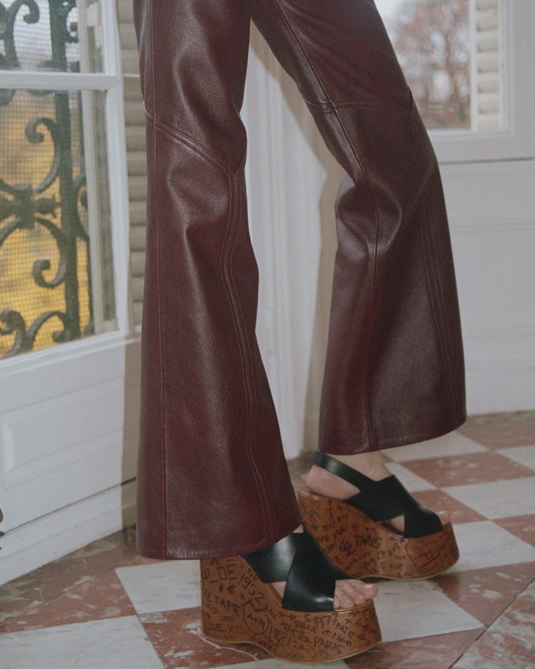 Calça de couro marrom de cintura alta e boca larga, combinada com sandálias plataforma preta de madeira com escritos gravados. Look moderno e ousado, alinhado às tendências de moda de street style e anos 70. nova era da Chloé