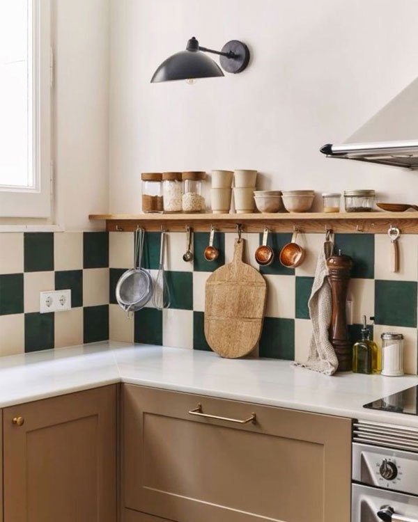 Cozinha moderna com armários bege, bancada branca e azulejos quadriculados verde escuro e branco. Prateleiras de madeira com potes e utensílios expostos. Decoração minimalista e funcional com influência escandinava.