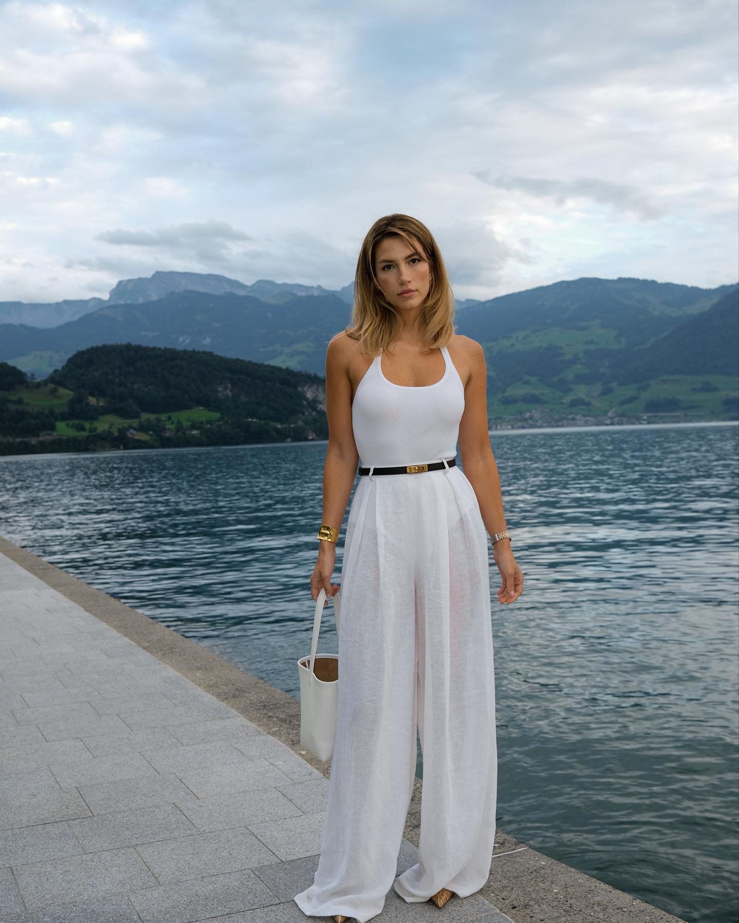 Mulher bem vestida com look elegante à beira de um lago. Ela usa uma regata branca, calça pantalona de tecido leve, cinto preto fino e acessórios dourados. Carrega uma bolsa grande branca.