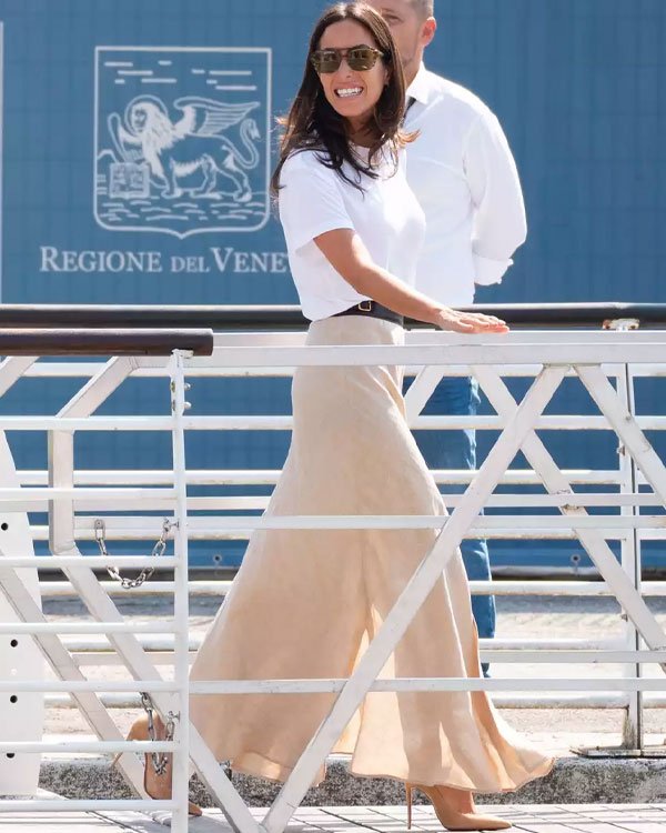 Mulher vestindo camiseta branca básica e saia longa bege, tendência casual e elegante. Complementa o look com óculos de sol aviador e sapatos de salto, representando uma moda prática e sofisticada.