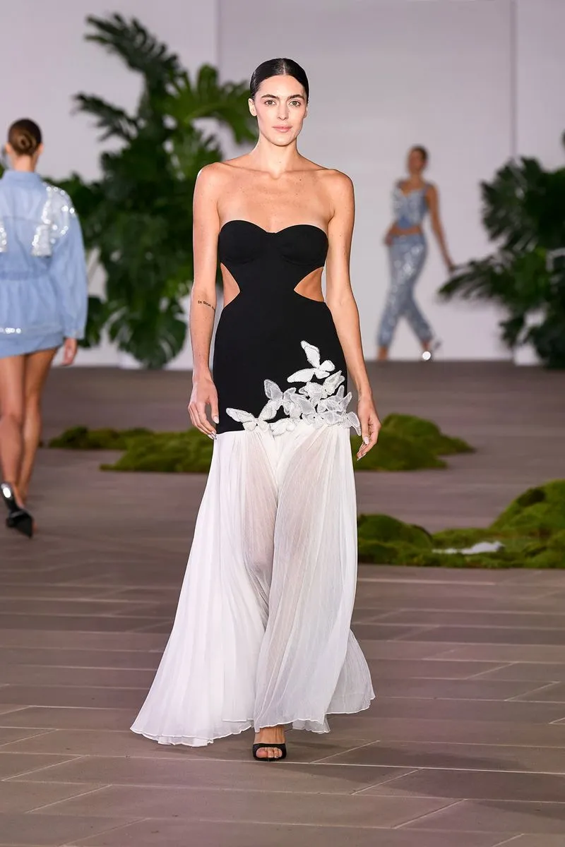 Modelo usa vestido longo preto e branco com recortes laterais e detalhes florais na passarela. O design combina elegância moderna e sofisticação, destacando tendências com recortes estratégicos e contrastes de cores. Estilo ideal para eventos formais.