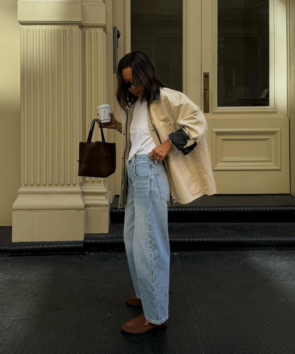 Mulher vestindo t-shirt branca, camisa bege oversized, calça jeans folgada e sapatos marrons. Acessórios incluem bolsa marrom e óculos escuros. Look casual e moderno, ideal para o dia a dia, seguindo tendências de moda confortável e prática.