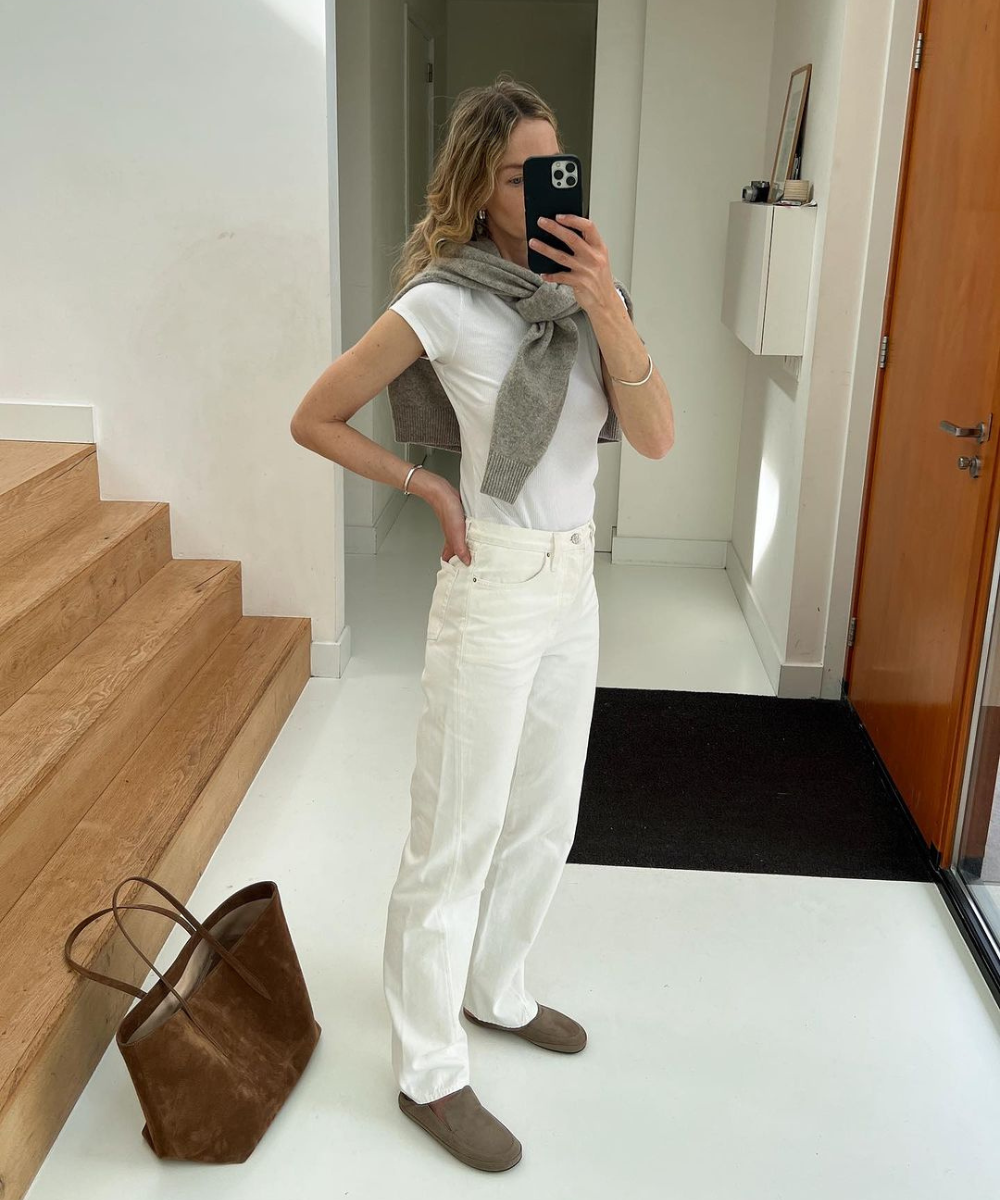 Look casual e minimalista: camiseta branca básica, calça jeans branca de corte reto, suéter cinza sobre os ombros, sapatos bege e bolsa de couro marrom. A combinação de cores neutras traz simplicidade e elegância ao estilo.