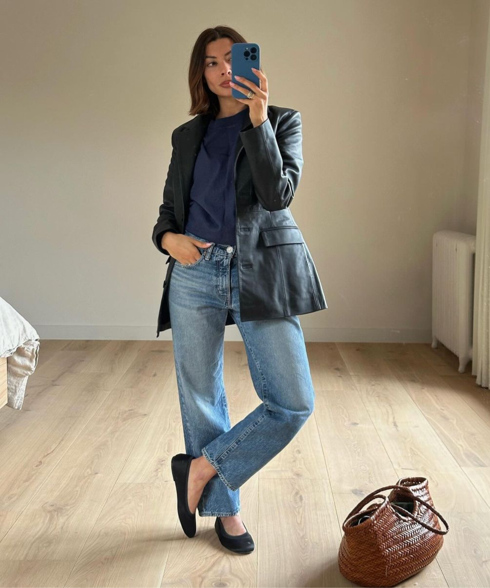 Mulher veste look casual chic, combinando blazer preto com camiseta azul marinho e jeans de cintura alta. Sapatos pretos e bolsa de palha completam o visual sóbrio e elegante. Tendência de moda mistura o clássico com o moderno, ideal para um dia a dia estiloso.