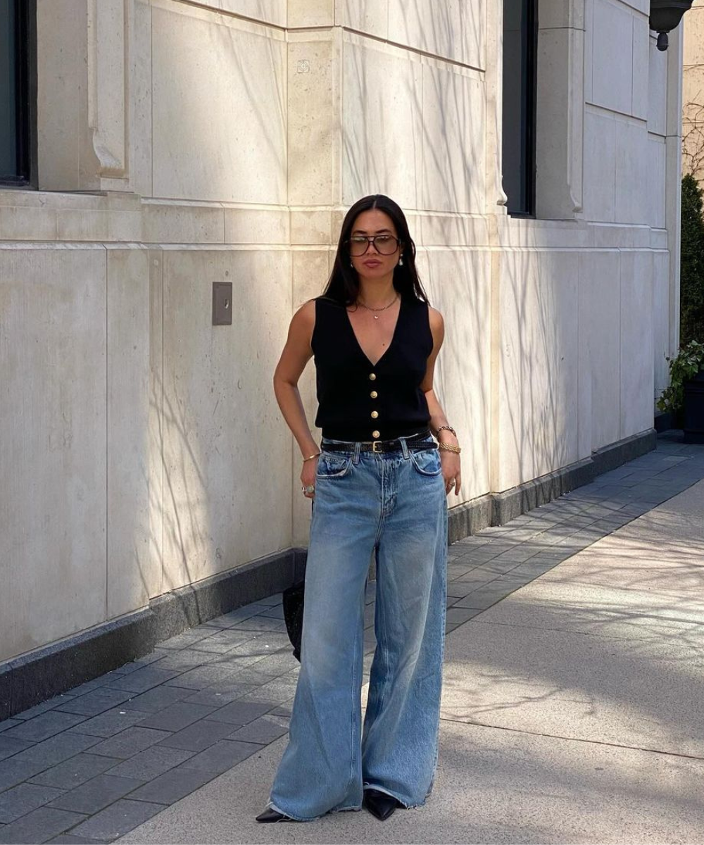 Mulher vestindo colete preto com botões dourados e calça jeans larga, estilo anos 90. Look casual e elegante, complementado por óculos de sol grandes e acessórios dourados. Tendência de moda urbana e descontraída.