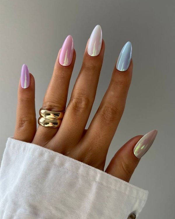 Unhas longas com esmalte em tonalidades pastel iridescentes, combinadas com anéis de metal dourado e camisa branca de tecido leve.