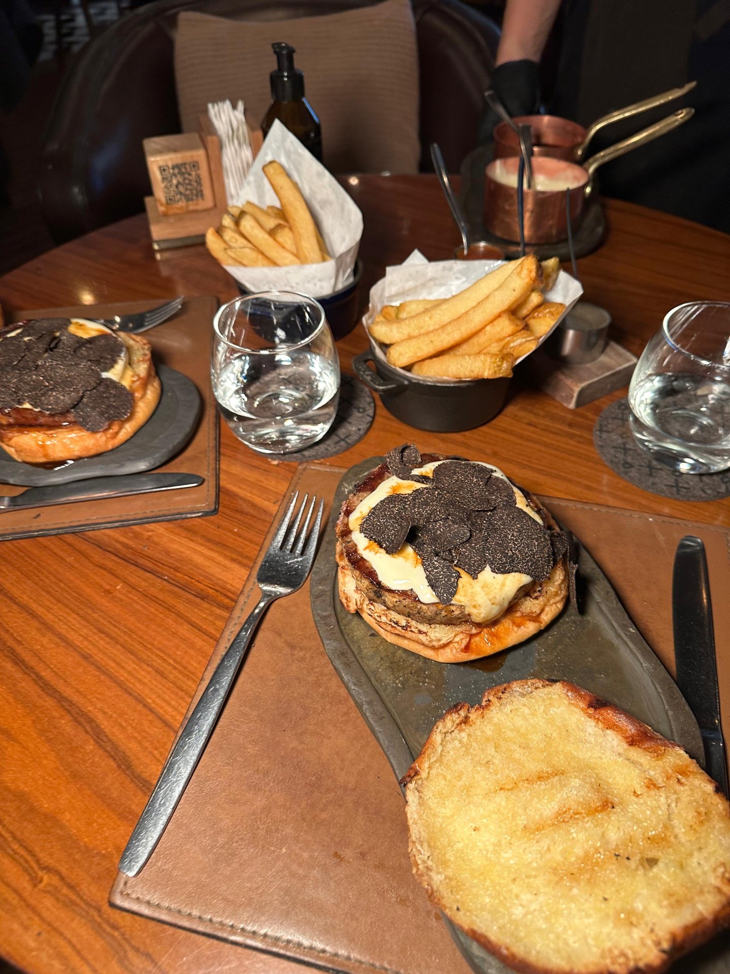 Mesa de restaurante sofisticado com hambúrgueres gourmet cobertos com trufas, acompanhados de batatas fritas grossas. Ambiente elegante com utensílios de cobre e prataria moderna. Restaurantes em Buenos Aires.