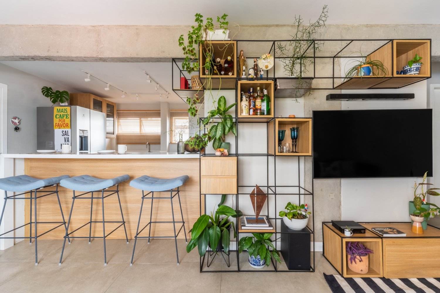 Cozinha e sala integradas com decoração moderna e minimalista. Estante metálica com nichos de madeira abriga plantas e objetos decorativos. Bancada com banquetas de design contemporâneo. Primeiro imóvel.