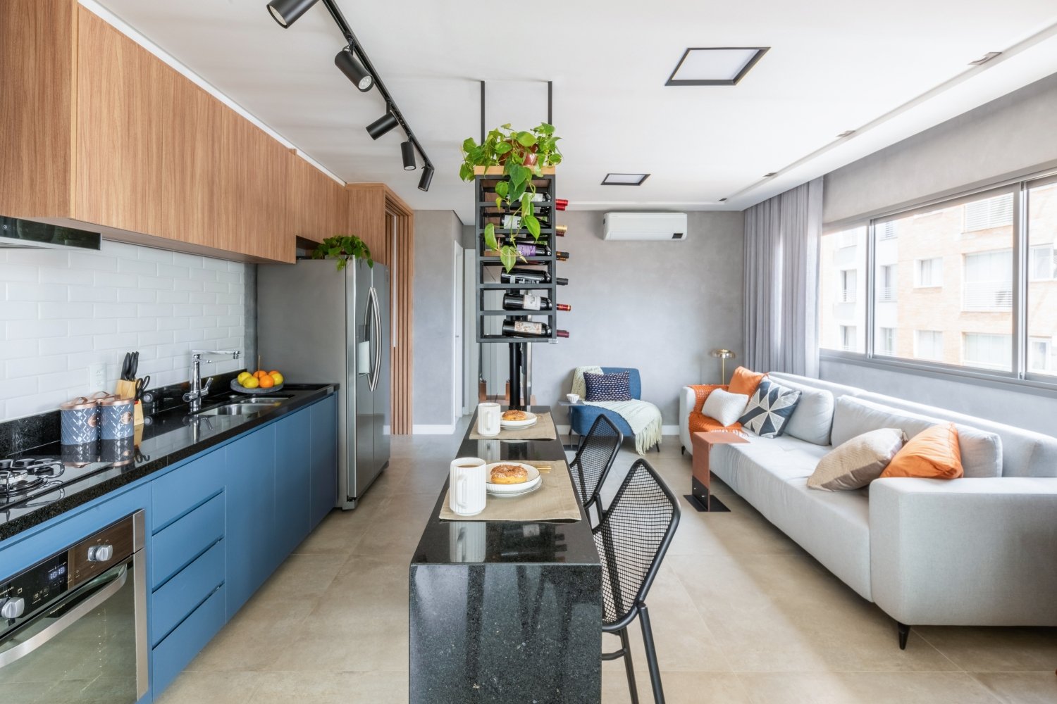 Apartamento moderno com decoração contemporânea. Cozinha integrada com armários azuis e de madeira, bancada preta e eletrodomésticos em inox. Sala de estar com sofá cinza claro e almofadas coloridas. Primeiro imóvel.