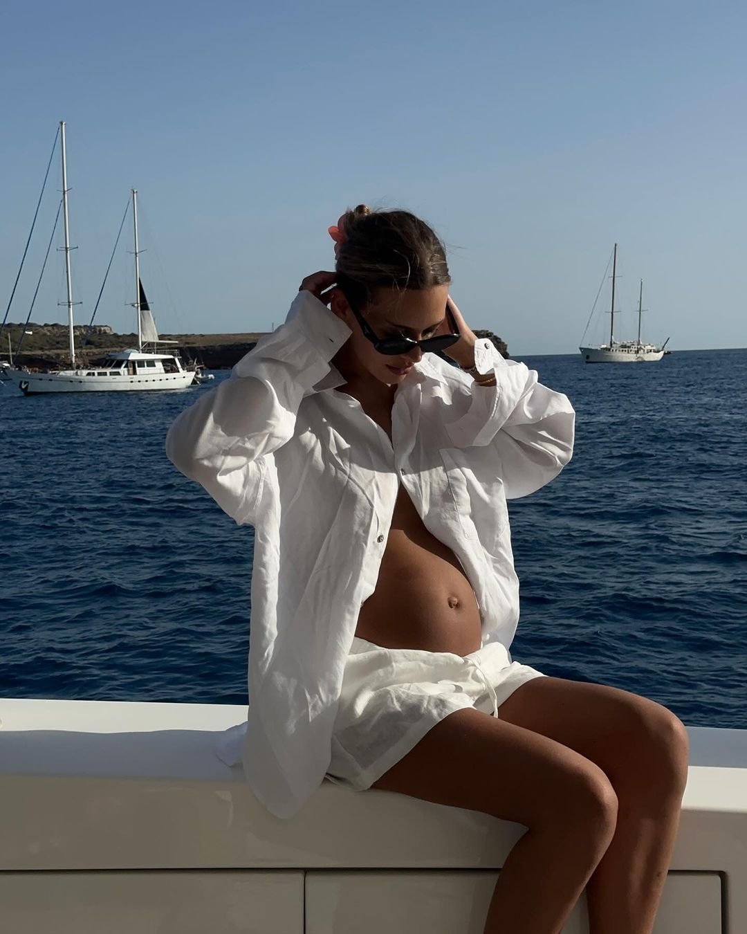 Mulher grávida em um barco, vestindo camisa e shorts brancos oversized, junto com óculos de sol modernos. O look combina conforto e estilo, destacando tendências de moda náutica e casual-chic. produtos super cheirosos